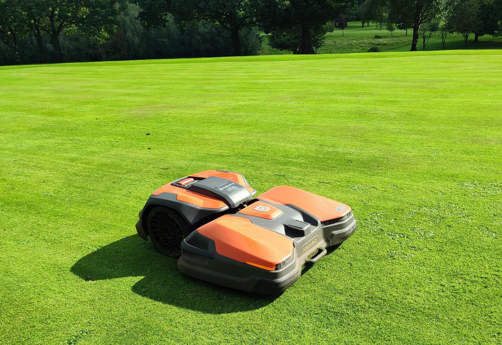 An orange and black robotic mower working on a golf course fairway in sunshine