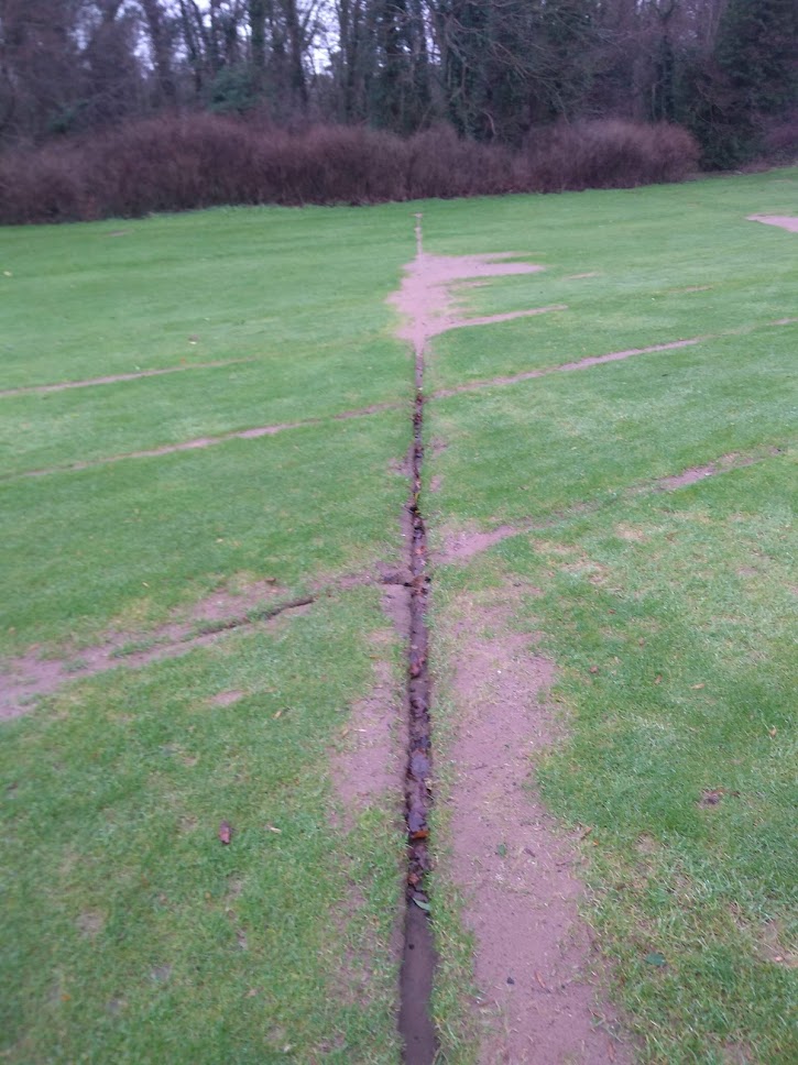 A gravel drain line running across a fairway, exposed at the surface