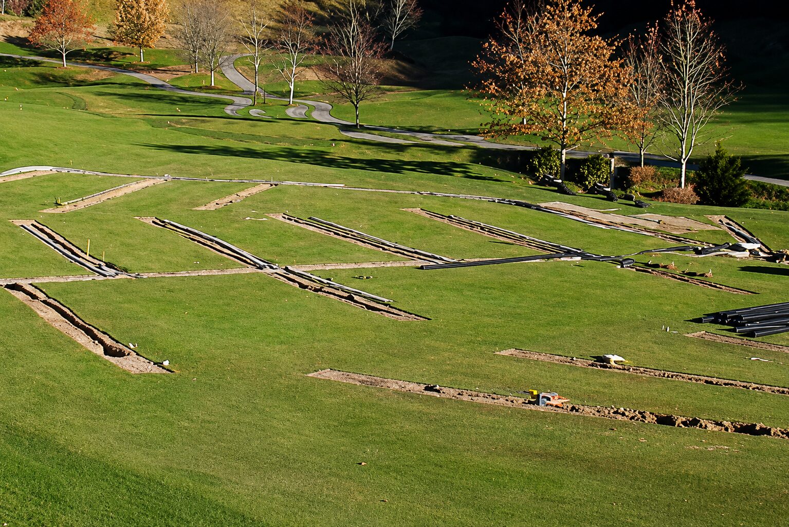 Aerial view of an extensive new drainage pipe network being installed across a fairway, with dozens of trenches and black pipe runs converging toward a collection point
