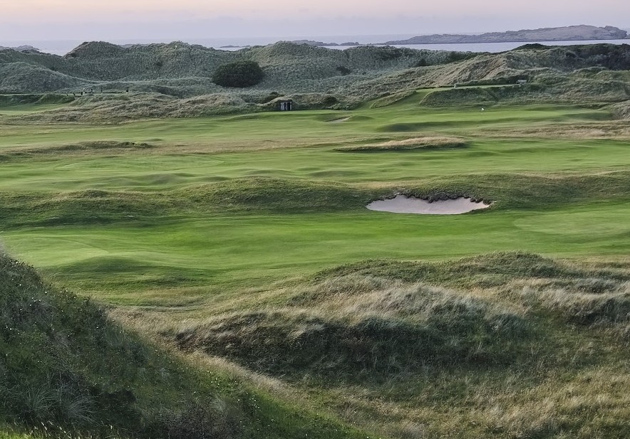 A natural links course at dusk, with dune grasses and gorse framing firm, undulating fairways and greens under a soft evening sky